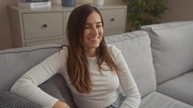 Genç bir kadın rahat bir oturma odasında rahat bir kanepede rahatlıyor, neşeyle gülümsüyor, evde huzurlu bir anın tadını çıkarıyor..