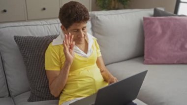 Sarı tişörtlü yaşlı İspanyol kadın evdeki oturma odasındaki kanepede dizüstü bilgisayarından video görüşmesi yapıyor.