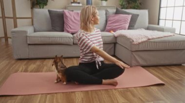 Genç sarışın bir kadın, oturma odasında pembe bir paspasın üzerinde yoga yapıyor. Yanında küçük bir chihuahua köpeği var..