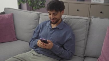 Genç, neşeli Arap adam evde bir kanepede oturuyor, akıllı telefon kullanıyor ve işaret dilini parmaklarıyla gösteriyor, tek gözüyle işaret ediyor..