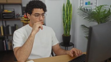 Ofis ortamında gözlük takan İspanyol bir adam arka planda kitaplıklar ve bitkilerle bir masada çalışırken atıştırmalık yiyor..