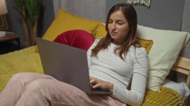 Sarı yastıkları, beyaz lambası ve arka planında bitkisi olan rahat bir yatak odasında dizüstü bilgisayarda çalışan genç, çekici bir kadın..