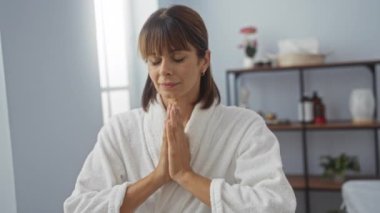 Spa odasında meditasyon yapan bir kadın beyaz bornoz giyiyor, gözleri kapalı, bir sağlık merkezinde huzuru ve arka planda bitkileri hissediyor.