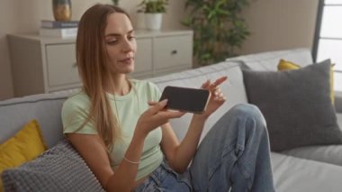 Etkileyici genç bir kadın evinde rahat bir oturma odasında otururken iletişim kurmak için akıllı telefon kullanıyor..