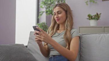 Modern bir oturma odasındaki koltukta akıllı telefon kullanarak gülümseyen genç sarışın kadın..