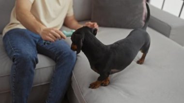 Gri bir kanepede oturan bir adam rahat bir oturma odasında bir köpekle oynuyor, yetişkin bir erkekle evcil hayvanı arasındaki bağı vurguluyor..
