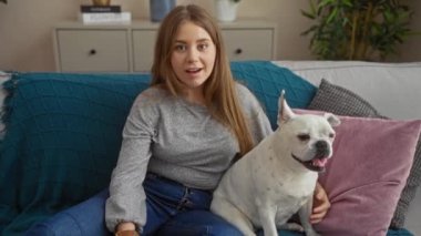 Neşeli, sarışın bir kadın evdeki rahat bir oturma odasında sevimli köpek evcil köpeğiyle kanepede oturuyor..