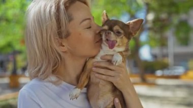 Sarışın genç bir kadın güneşli bir günde şehir parkında köpeğini sevgiyle kucaklıyor ve öpüyor..