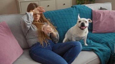 Genç sarışın bir kadın, elinde fincanla alnına dokunuyor, rahat bir oturma odasında, mutlu beyaz bir köpekle koltukta oturuyor..