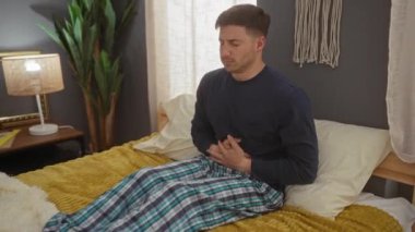 Pijamalı genç bir adam yatak odasındaki bir yatağa oturur, aşağı bakar ve midesini rahatsız bir şekilde komodinin yanında bir lamba ve bir bitkiyle tutar..