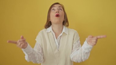 Şaşırmış orta yaşlı İspanyol kadın, şaşırmış bir şekilde ayakta duruyor, izole edilmiş sarı arka plana bakıyor.