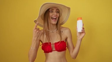 Bikinili beyaz bir kadın güneş kremi tutuyor ve izole edilmiş sarı arka planda mutlu bir şekilde gülümsüyor.