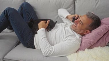 Orta yaşlı bir adam, rahat bir oturma odasında iki Chihuahua ile koltukta dinleniyor telefonda konuşurken gülümsüyor..