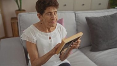 Beyaz bluz giyen kısa saçlı yaşlı İspanyol kadın oturma odasında oturuyor, çerçeveli bir fotoğrafı yakından inceliyor ve gri bir kanepede minderlerle oturuyor..