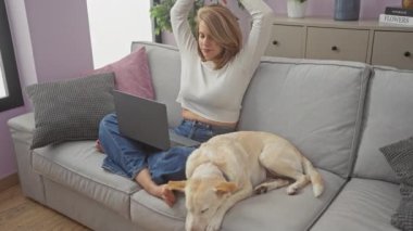 Genç bir kadın, rahat bir oturma odasındaki kanepede uyuyan köpeğinin yanında dizüstü bilgisayarla çalışırken esniyor..