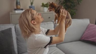 Sarışın, beyaz bir kadın oturma odasında Chihuahua köpeğini tutuyor, sıcak bir kanepede, modern bir ev ortamında sevgi dolu bir anı paylaşıyor..