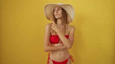 Bikini ve yaz şapkalı beyaz kadın, izole edilmiş sarı arka planda parmakla işaret ediyor, ciddi bir yüz ifadesi var.