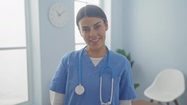 Önlüklü, steteskoplu, gülümseyen genç bir kadın, parlak bir hastane odasında duruyor..