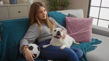 Sarı saçlı genç bir kadın oturma odasında bir kanepede oturuyor, elinde bir futbol topu tutuyor ve beyaz bir köpeğe sarılıyor..