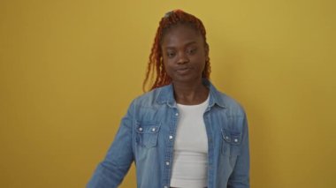 Kot gömlek giyen neşeli Afro-Amerikalı kadın, izole edilmiş sarı bir arka plan karşısında duruyor, gülümsüyor ve zafer sembolünü yüzünde parmaklarıyla gösteriyor.