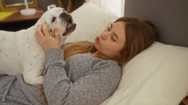 Genç sarışın bir kadın yatak odasındaki bir yatakta beyaz köpeğiyle oynarken sıcak ve rahat bir yuva kuruyor..