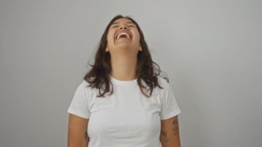 Ayakta duran ve gülen bir kadın, beyaz tişört giyen genç bir İspanyol kadın, izole bir arka planda yüksek sesle gülümsüyor, komik bir şaka yüzünden, eğleniyor ve yüksek sesle kahkaha atıyor.