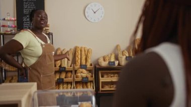 Fırında müşteriyle konuşan bir kadın, arka planda raflar dolusu ekmekle duvardaki saatin altında.