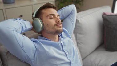 Genç, yakışıklı, sakallı, kulaklıkla müzik dinleyen, rahat bir dairenin oturma odasındaki koltukta dinlenen, mavi gömlekli, evde oturan bir adam..