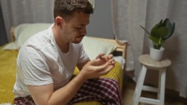 Yatak odasındaki genç İspanyol bir adam sarı bir battaniyeyle kaplı bir yatakta oturuyor. Ellerini düşünceli ve düşünceli bir şekilde yüzünü örtüyor. Yanında yeşil bir bitkisi olan küçük beyaz bir tabure var..