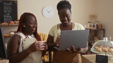 Afro-Amerikalı kadınlar fırın odasında gülümserken dizüstü bilgisayar kullanıyorlar ve sohbet ediyorlar..