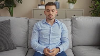 Latin kökenli genç bir adam, rahat bir oturma odasında, açık mavi gömlekli, ev bitkileri ve dekorlarla çevrili, düşünceli ve rahat görünüyor..