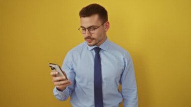 İspanyol asıllı genç adam tişört ve kravat giyerek mutlu bir şekilde dilini dışarı çıkarırken akıllı telefonu izole edilmiş sarı arka plan üzerinde kullanıyor. Eğlenceli ve eğlenceli ifadeler.!