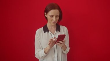 Kafkasyalı genç bir kadın, izole edilmiş kırmızı arka planda duruyor. Öfkesini ve öfkesini çılgınca bağırışlar ve bağırışlarla ifade ediyor. Sinirli ve sinirli bir şekilde akıllı telefonunu kullanıyor.