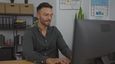 Modern bir ofis odasında, masaüstü bilgisayarında çalışan genç İspanyol bir adam elleri suratında stresli görünüyor.