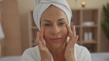 Orta yaşlı bir kadın kapalı bir yerde spa tedavisinden zevk alırken dingin bir salon ortamında cilt bakım ürünleri uyguluyor.