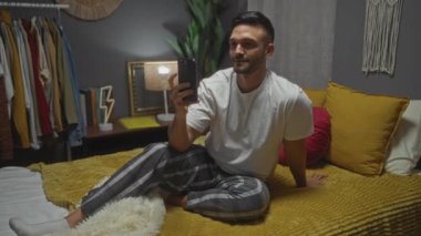 Yakışıklı bir İspanyol adam şık ve şık bir yatak odasında yatağında otururken akıllı telefonuyla selfie çekiyor..