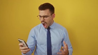 Gömlekli ve kravatlı öfkeli genç adam akıllı telefona bağırıyor, öfkeden kuduruyor, sarı arka planda izole edilmiş.