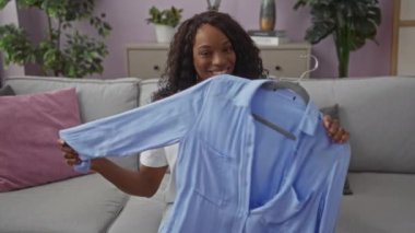 Güzel, genç, Afro-Amerikalı, kıvırcık saçlı, gülümseyen, mavi gömlekli, rahat bir oturma odasında kanepede oturan bir kadın..