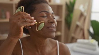 Orta yaşlı bir kadın, yumuşak doğal ışık ve sakin bir ortam ile sakin bir spa ortamında yeşim taşıyla yüz masajından zevk alıyor.