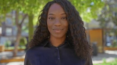 Kıvırcık saçlı güzel, Afro-Amerikan bir kadın şehir parkında gülümsüyor..