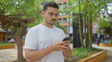 Güneşli bir günde şehir parkında akıllı telefon kullanan genç bir İspanyol açık havada modern bir yaşam tarzı sergiliyor..