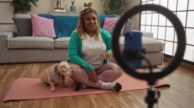 Saçları örgülü genç bir Afrikalı Amerikalı kadın oturma odasında köpeğiyle birlikte yoga minderinde oturup video kaydediyor..