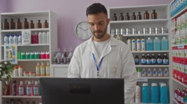 Sakallı, genç, yakışıklı bir adam bir eczanede beyaz önlük giymiş ve tezgahın arkasında çalışıyor. Arka planda çeşitli ilaç şişeleri ve ürünleri var..