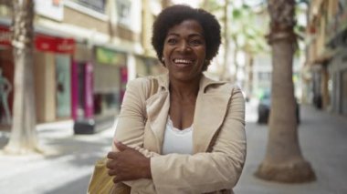 Kıvırcık saçlı orta yaşlı bir kadın, bej bir ceket giyiyor, dükkanları ve palmiyeleri olan bir sokakta gülümsüyor, güvenini kollarını kavuşturarak ifade ediyor.