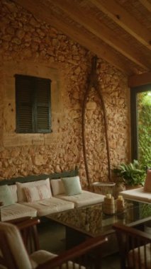 Konforlu oturma odası ve yastıklarla ve asma kaplı pencerenin yakınındaki ahşap tavanın altında ahşap elementlerle aksanlı cam sehpası olan kapalı taştan bir yaşam alanı.