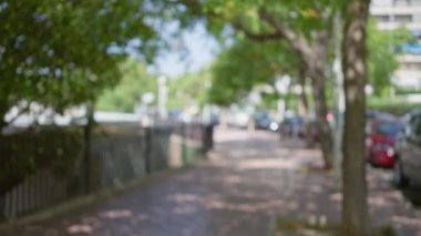 Bulanık kaldırımda ağaçlar ve park halindeki arabalarla gündüz vakti gezen kişi.