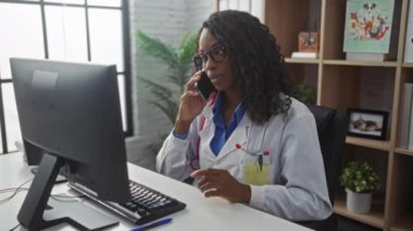 Kıvırcık saçlı, gözlüklü, bilgisayar başında çalışan ve modern bir klinikte telefonla konuşan Afrikalı Amerikalı kadın doktor..