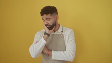 Genç İspanyol bir adam, önlük giymiş, yorgun ve sıkılmış görünüyor, izole edilmiş sarı bir arka planda sorunlarını düşünüyor, mücadele ve depresyon tasviri.