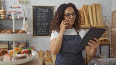 Fırında çalışan bir kadın, tablet kullanırken telefonla konuşuyor. Etrafı ekmek, pasta ve tatlı ile çevrili..