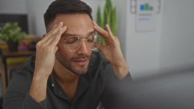 Odaklanmış, sakallı, gözlüklü, ofis ortamında başını kapalı tutan, arka planda bitki ve çizelgeleri olan İspanyol bir adam..